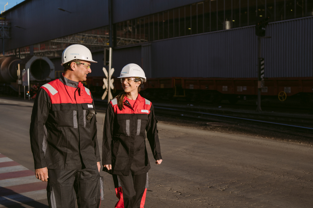 Mitarbeiterin und Mitarbeiter, Vater und Dochter mit Arbeitskleidung und Helm im Werksgelände der voestalpine Donawitz