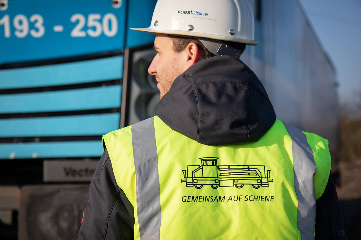 Benjamin von hinten mit der Werksbahn Weste mit der Aufschrift Gemeinsam auf Schiene vor einer Lok