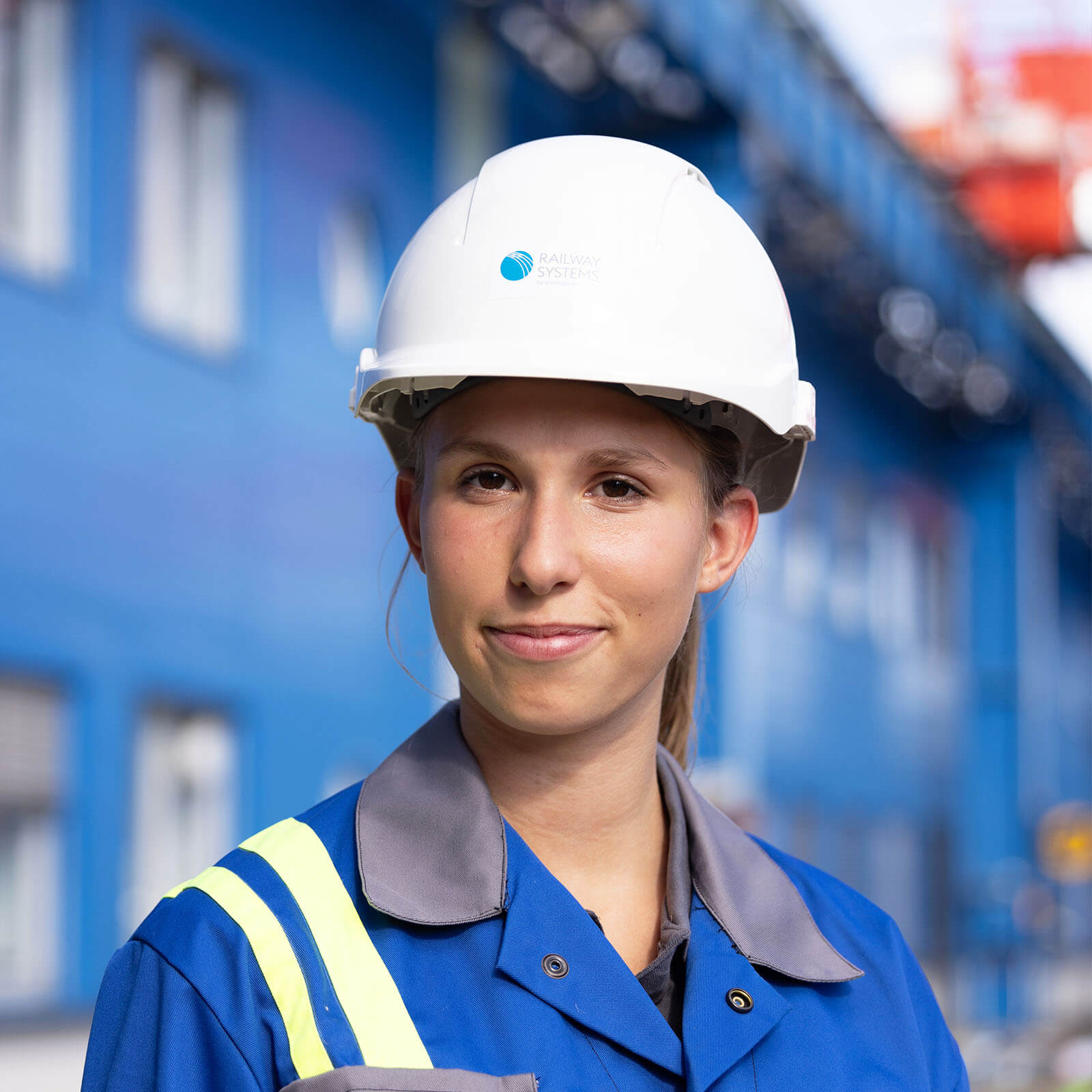 voestalpine employee Lisa looks into the camera with a white hard hat and blue work clothes, in the background you can see an industrial area.