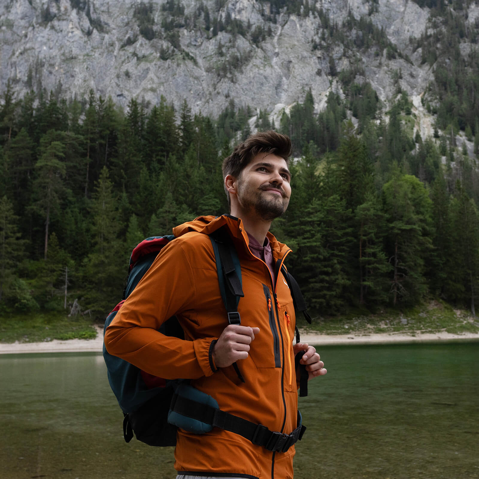 Voestalpine employee Michael stands in his hiking gear with a rucksack in front of a steep mountain slope