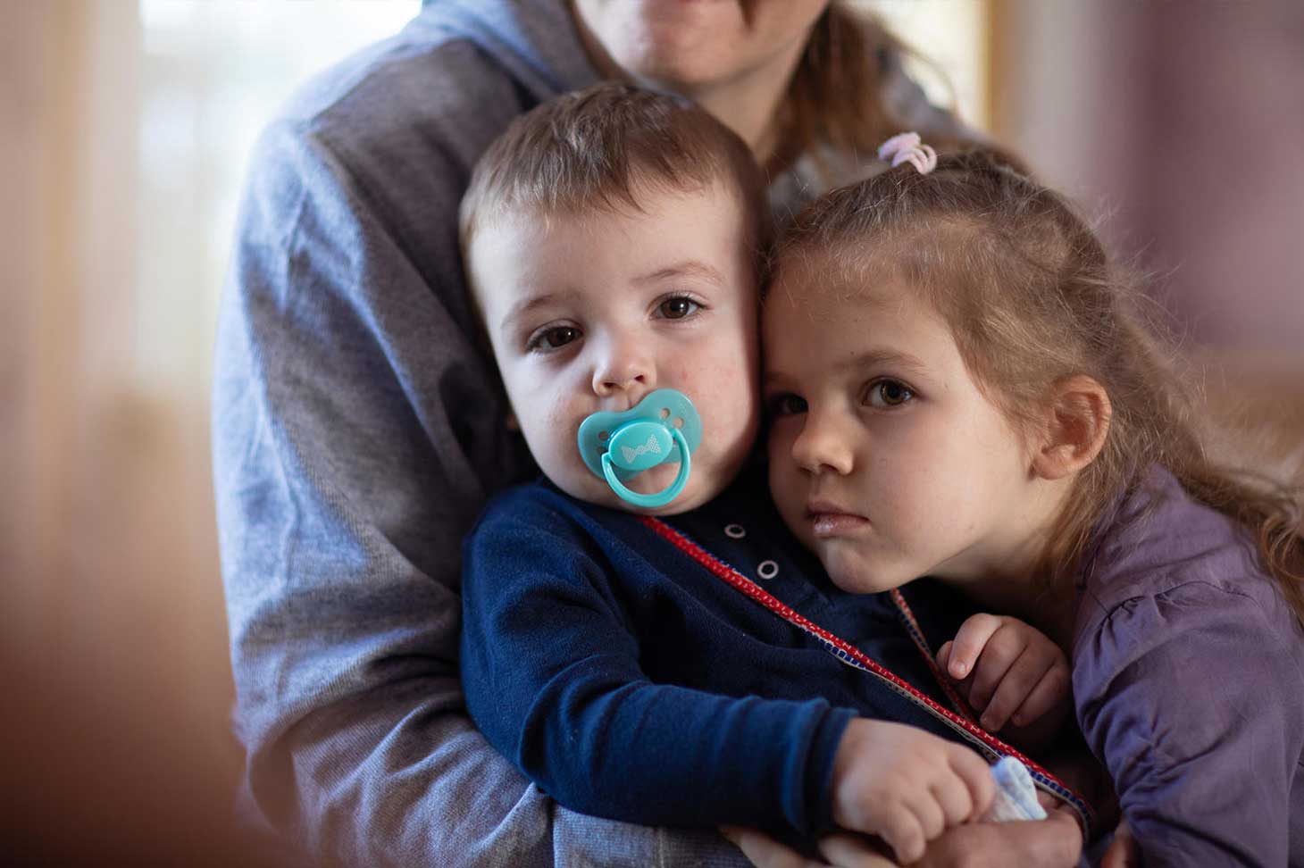 A toddler sits on an adult's lap with a pacifier in her mouth, a girl leans her forehead against his cheek, both look into the camera.