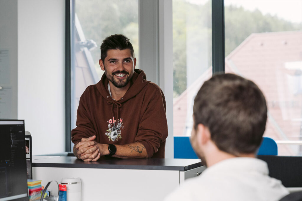 Michael is leaning on a desk talking to a colleague