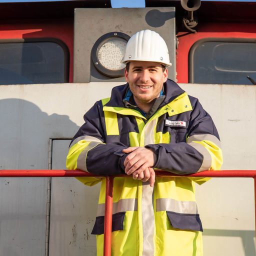 Benjamin in seiner Arbeitskleidung und mit weißem Helm auf einer Werksbahnlok der LogServ