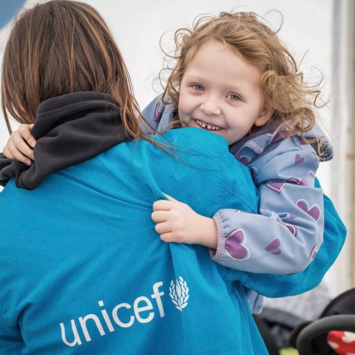 Ein Mädchen lacht in die Kamera und wird gehalten von einer unicef Mitarbeiterin, die mit dem Rücken zur Kamera steht