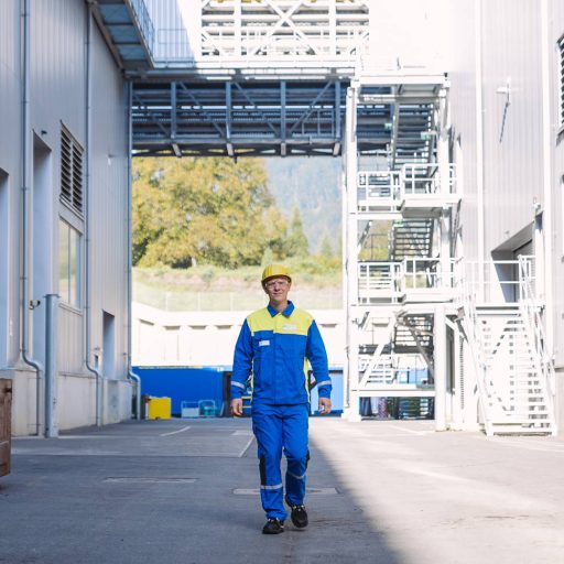 voestalpine Mitarbeiter Christoph geht in seiner Arbeits-Schutzbekleidung zwischen zwei Werkshallen auf die Kamera zu