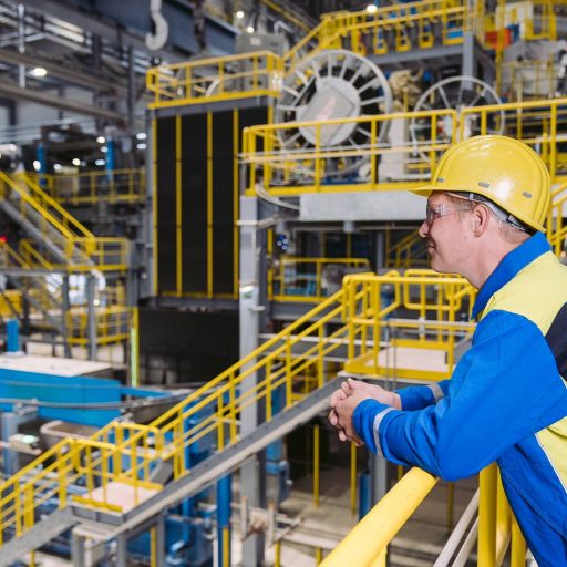 voestalpine Mitarbeiter Christoph lehnt in seiner Schutzkleidung mit Sicherheitshelm am Geländer einer voestalpine Werkshalle und blickt Richtung links