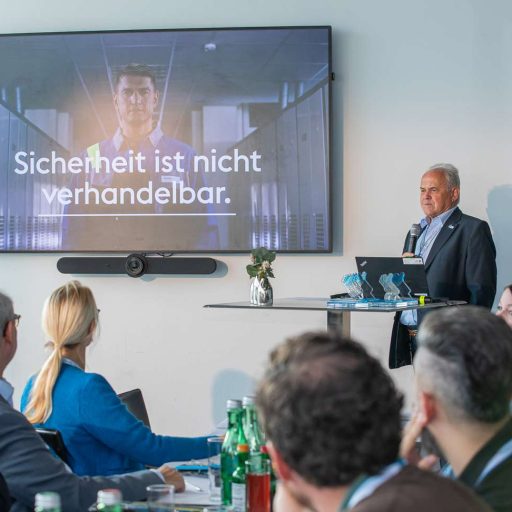 People sit around a table and follow a presentation by a man with a microphone on the subject of safety at voestalpine. The screen reads Safety is non-negotiable