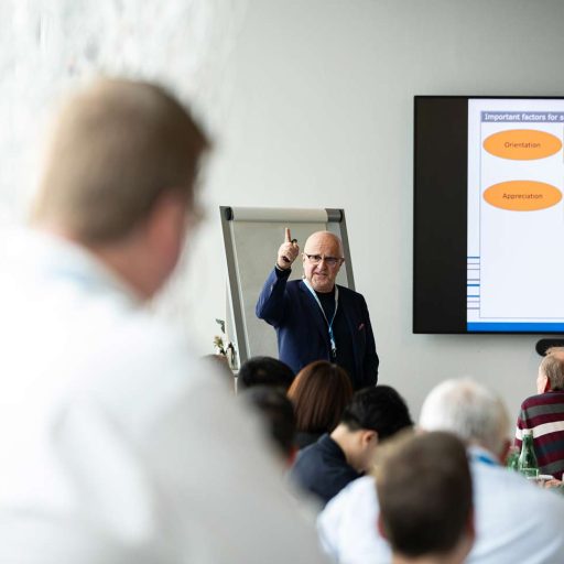 A lecturer points his index finger upwards while giving a voestalpine safety training course, which is followed by many people in the room.