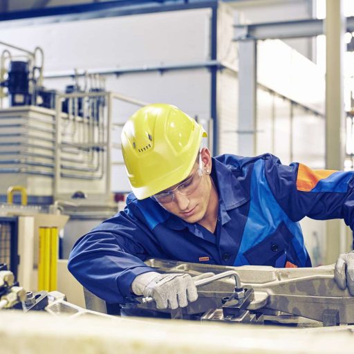 voestalpine worker wearing a hard hat and safety goggles screws a bolt into a steel part on a factory floor