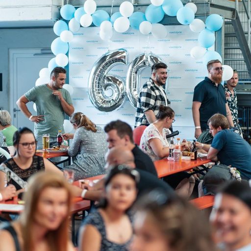 Gäste sitzen an Bierbänken in der Halle des Werks in Dettingen, ein 60-Lufballon weißt auf das Jubiläum hin.